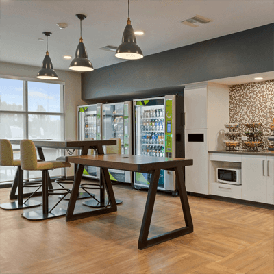 Lobby of an Extended Stay America hotel, showing two wooden dining tables, coolers with ready-to-go drinks, produce and bar seating.