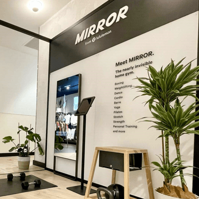 White wall with black accents and a large lcd screen showcasing the Lululemon MIRROR, a home gym. Black hand weights and potted plants surround.