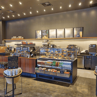 Front counter of a remodeled Starbucks coffee shop, with ready-to-eat snacks and beverages in chilled displays, warm lighting and earth-toned walls and flooring.