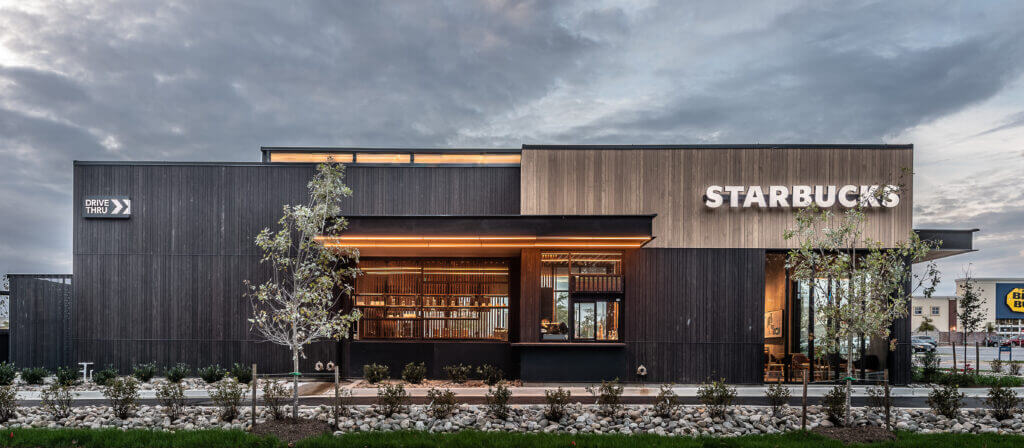 Front entrance of a newly remodeled Starbucks coffee shop, with wooden exterior, backlit displays and fresh landscaping.