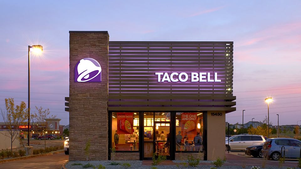 Night view of a Taco Bell restaurant, purple lights illuminating the storefront, with colorful signs in the windows advertising new menu items.