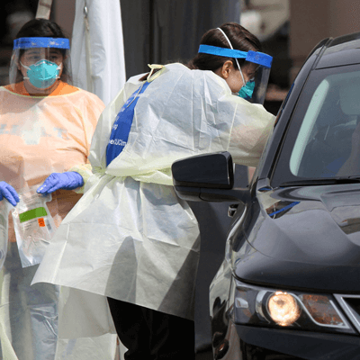 Nurse wearing white protective clothing and a blue face shield reaching into a black car to test a passenger for Covid-19.