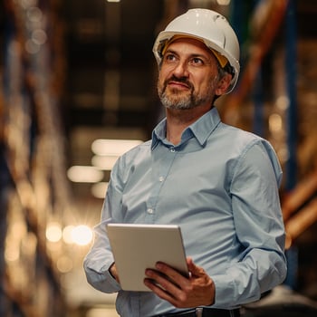 Man wearing a blue shirt and white hard hat, holding a clipboard in a warehouse